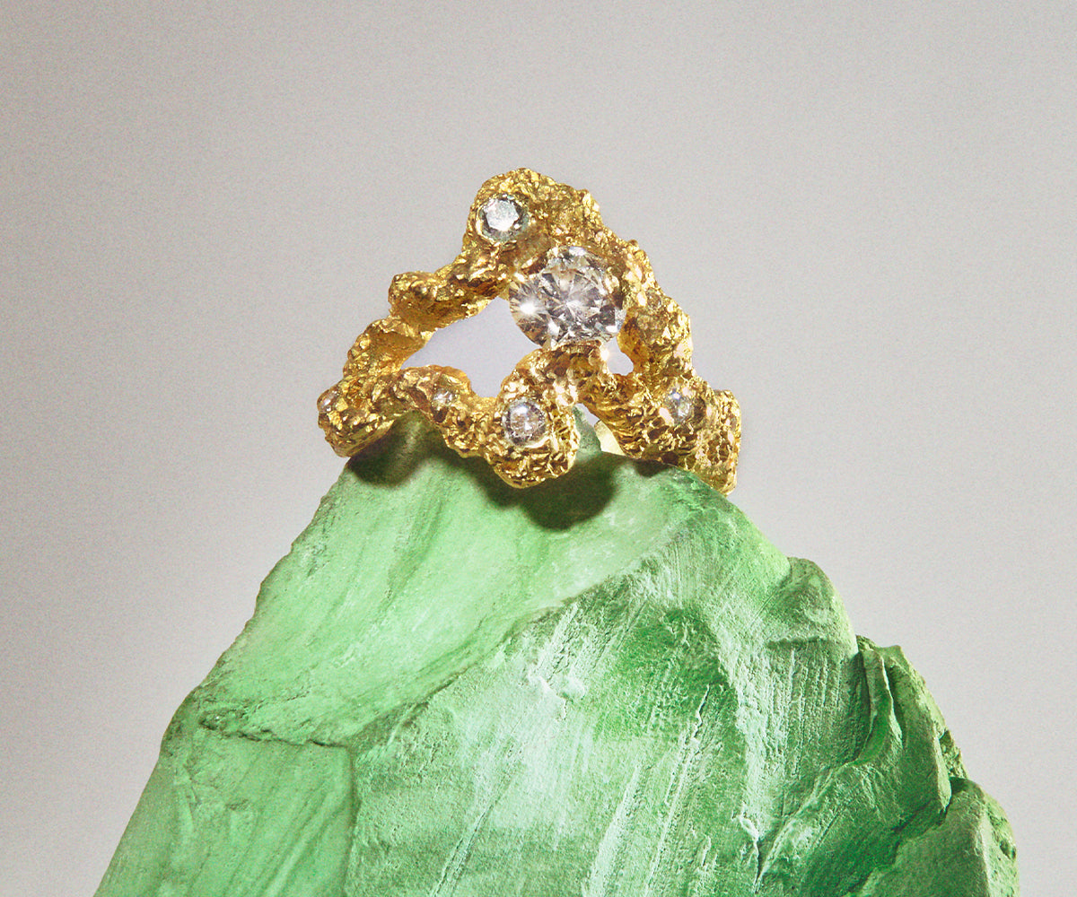 An 18K yellow gold ring with 13 small diamonds, shaped organically with a big stone centred in the middle. Placed on a green rock.