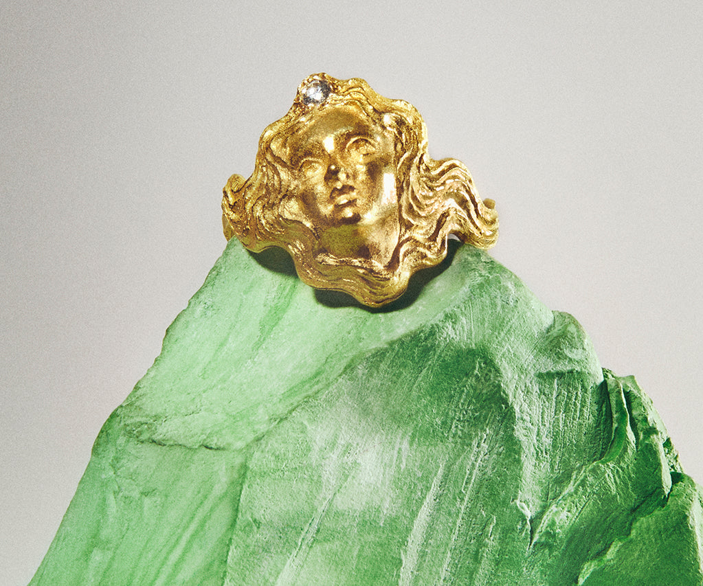 An 18K yellow gold ring resembling a small face, with a tiny diamond set in her hair, which forms the ring's shank.