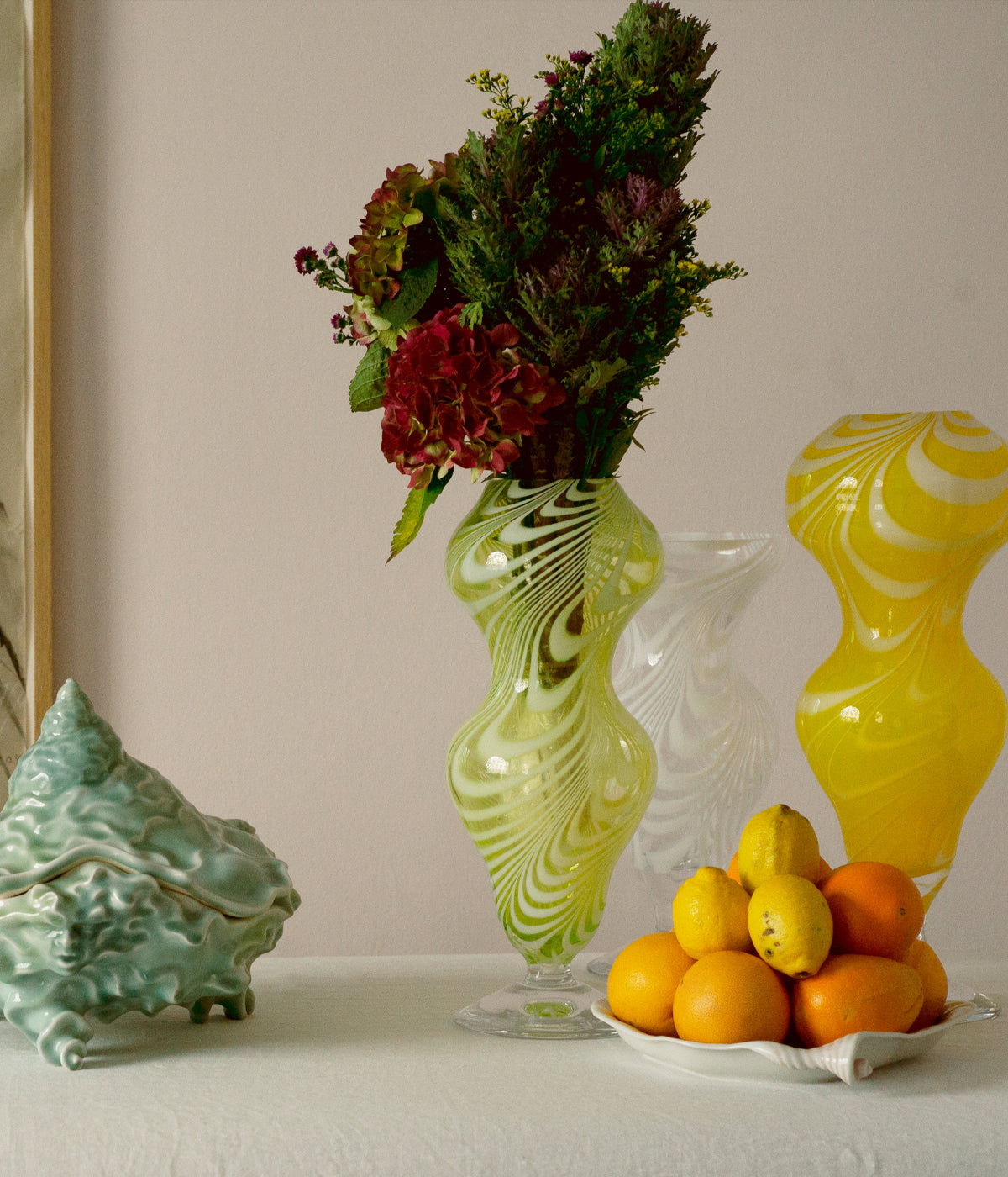 A table set with a tureen, oranges and 3 glass vases, one of them filled with flowers.