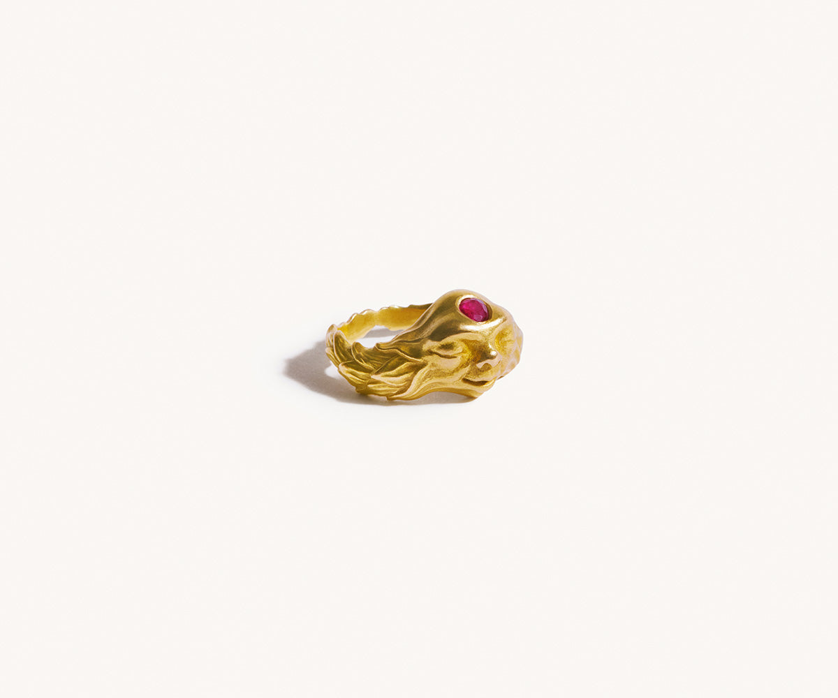A front-facing 18K yellow gold ring featuring a gnome with closed eyes and a red ruby on its forehead.