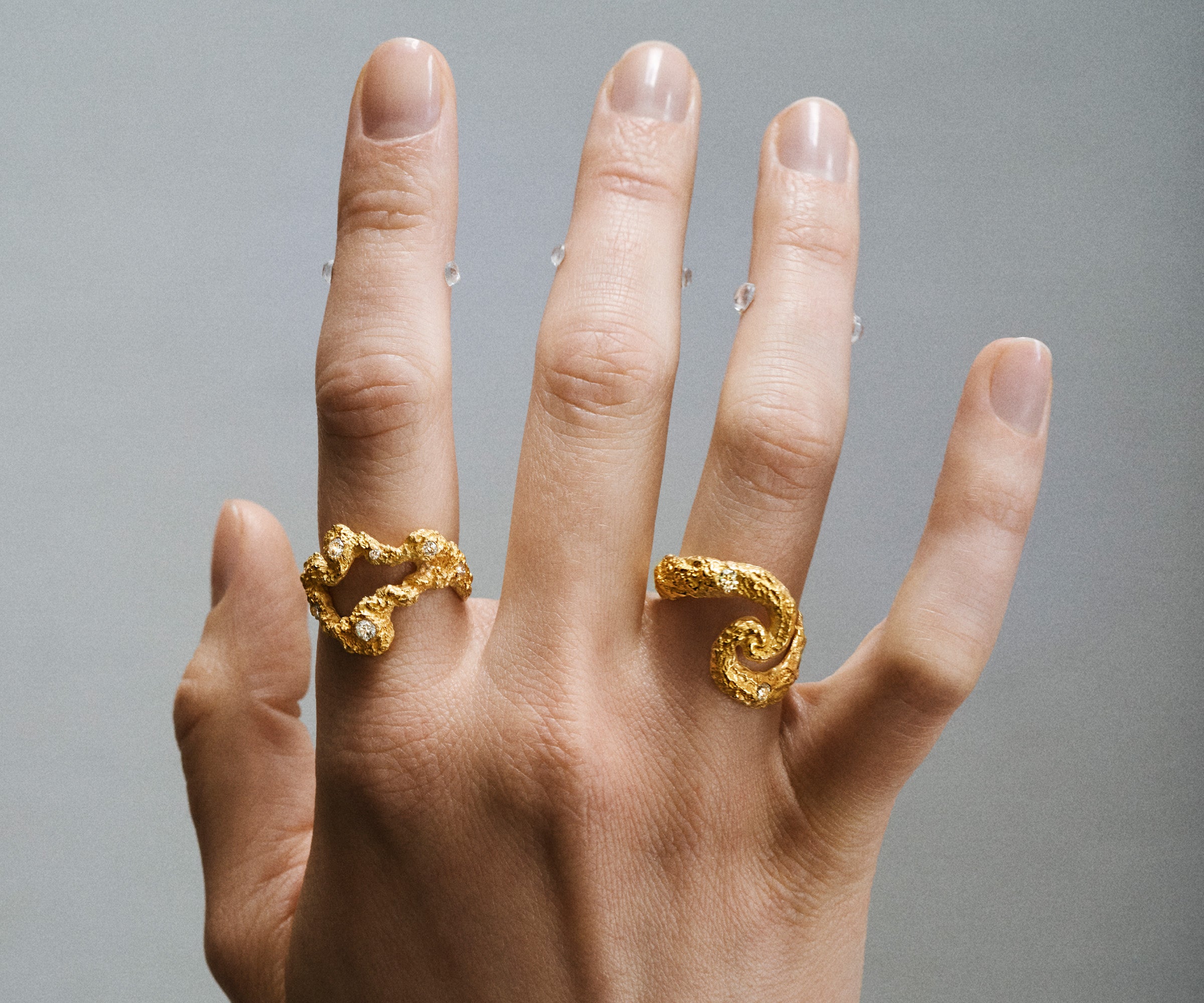 A hand with two 18K yellow old rings,  shaped organically with a whole in the middle in front and the other one like a 'S'. The surface of the gold resembles craters.