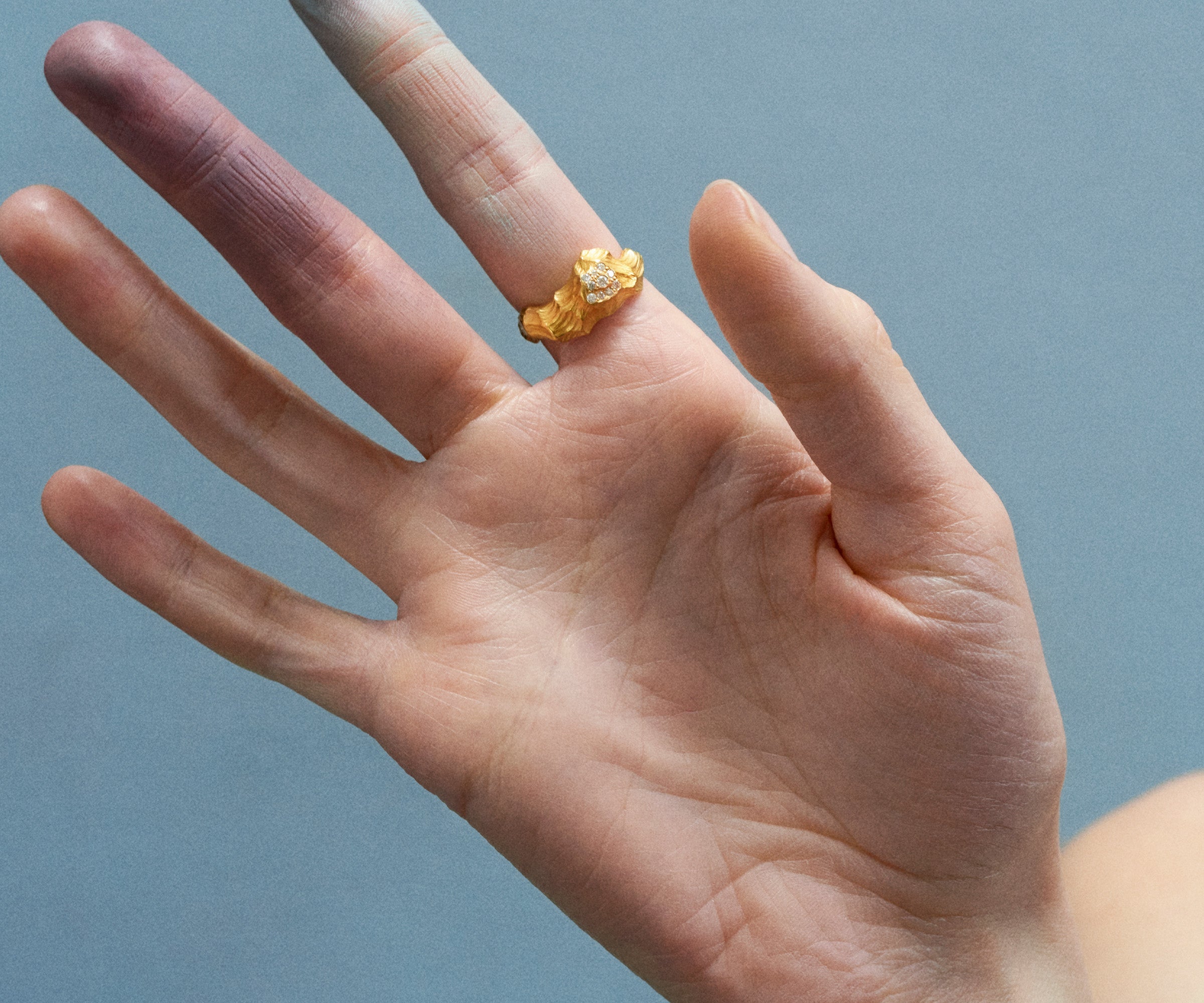 A hand wearing an 18K yellow gold ring shaped like waves in water, with 10 small diamonds set along the front.
