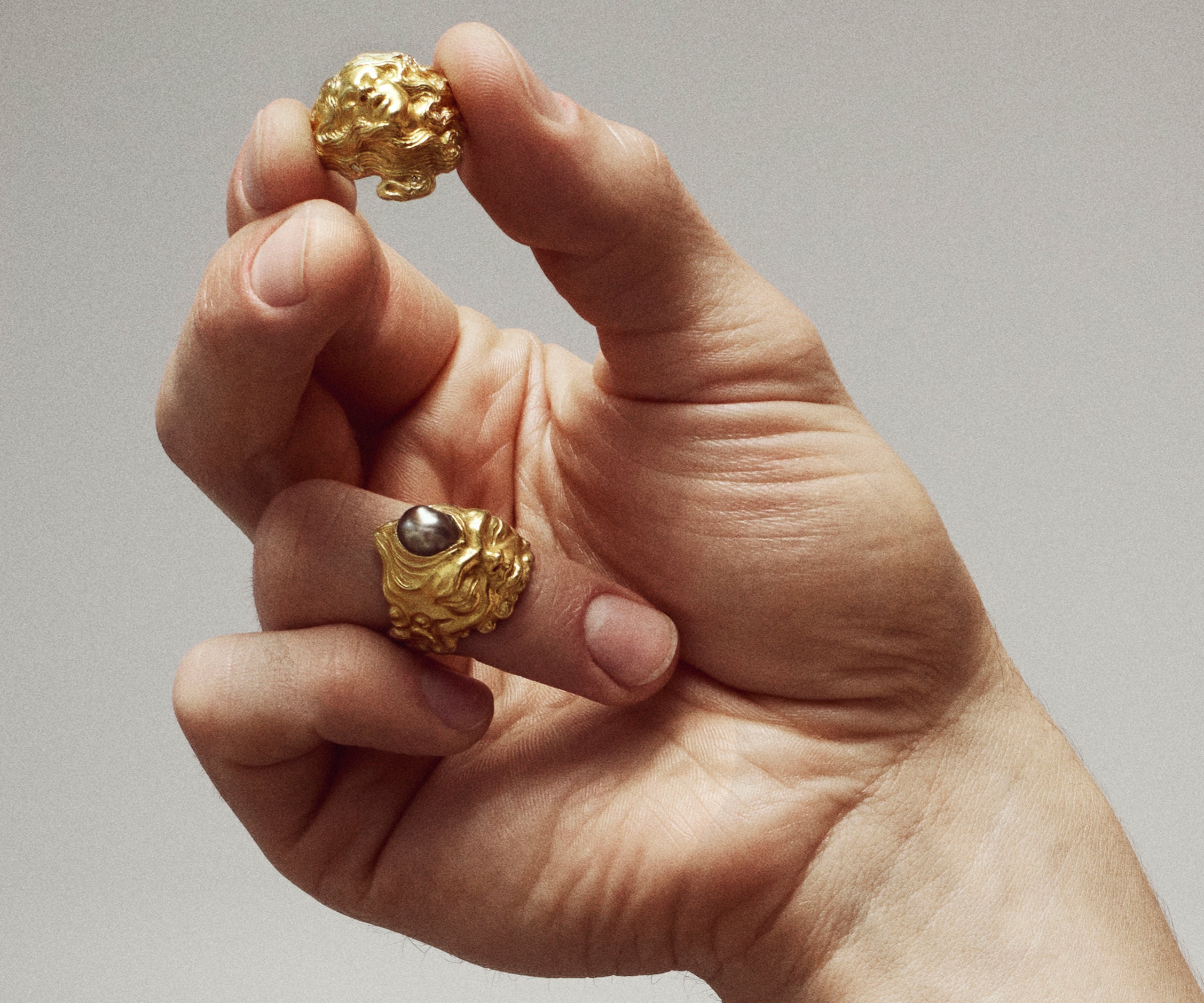 A male hand wearing an 18K yellow gold ring shaped like a gnome, with a black Tahitian pearl emerging from its forehead.
