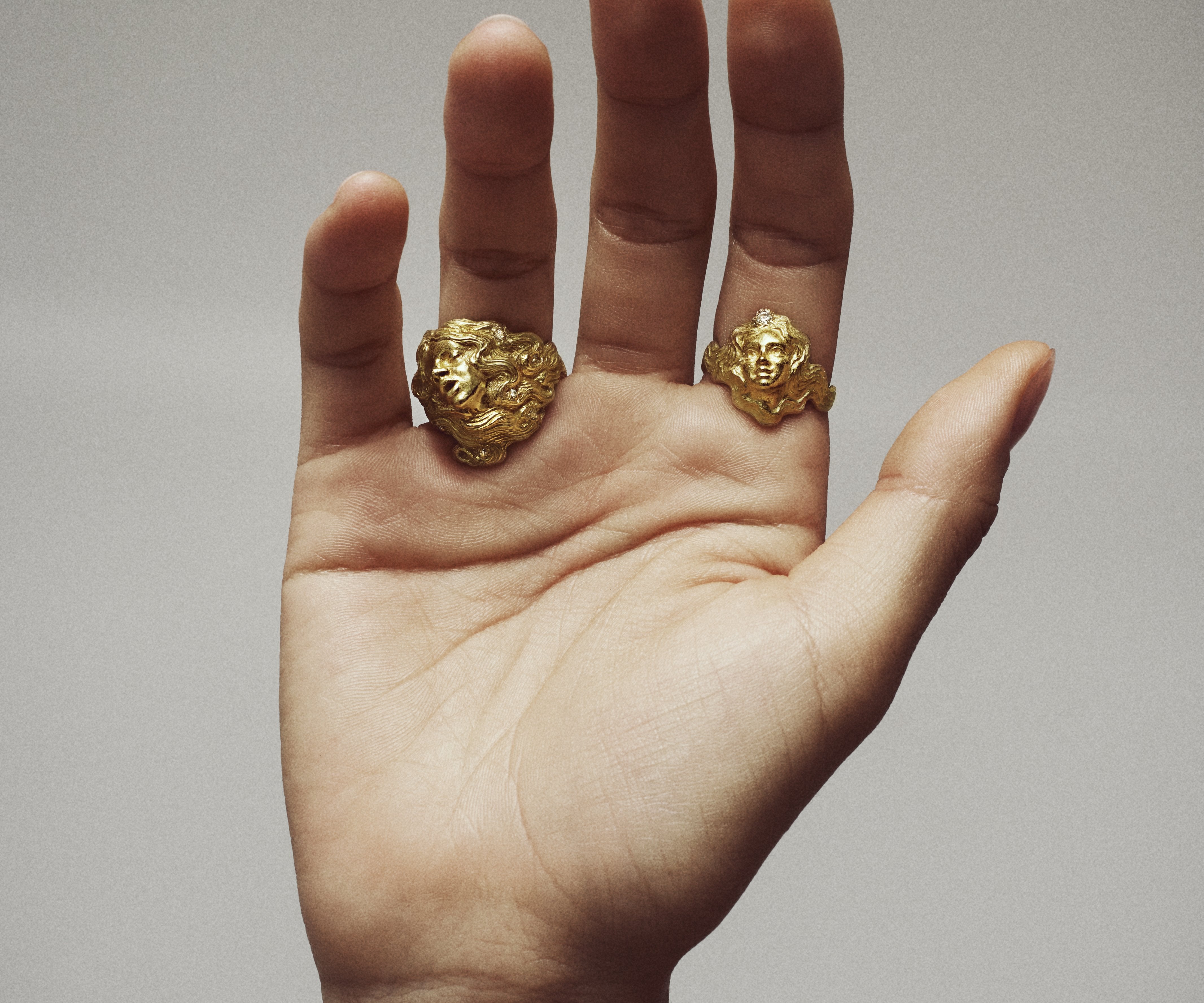 A hand wearing two rings, each resembling a woman emerging from the hair.